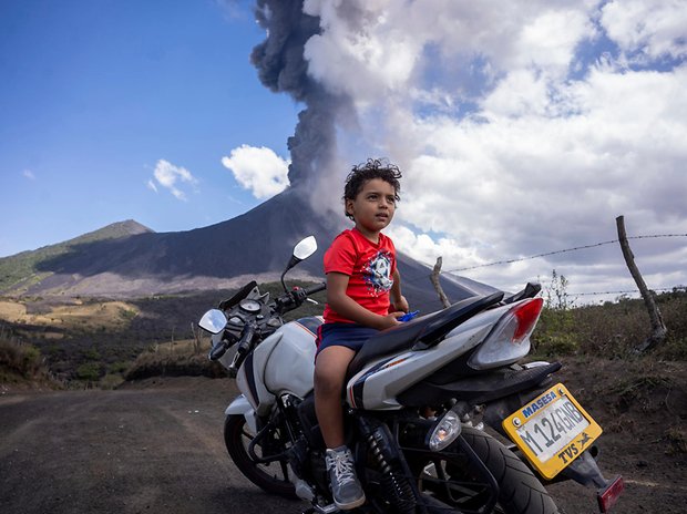 Ein Kind sitzt auf einem Motorrad, während im Hintergrund der Vulkan Pacaya ausbricht.