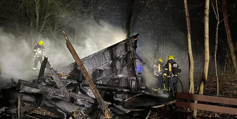 Feuerwehrleute löschen den brennenden und schon in sich zusammengefallenen Holzunterstand.