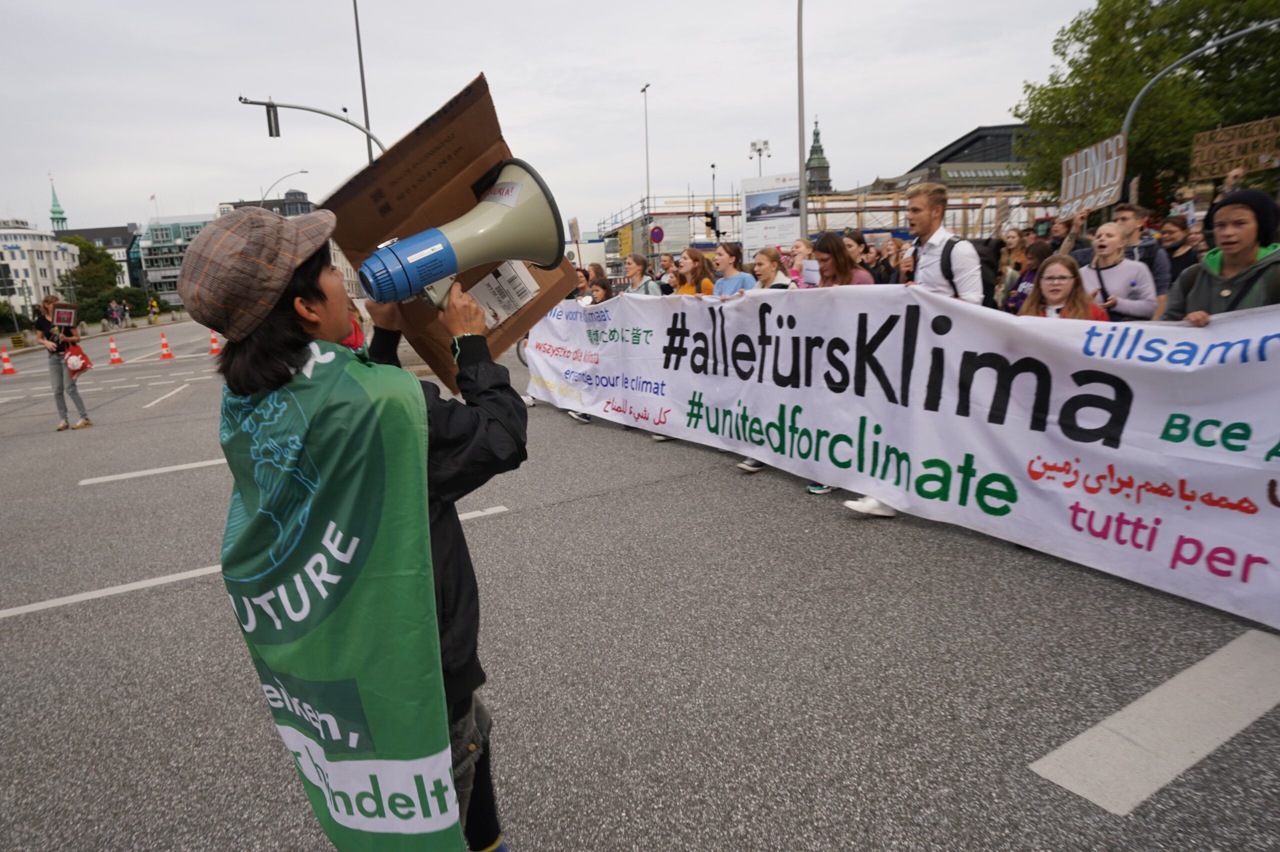 Ein Demonstrant hält ein Megaphon vor dem Mund und spricht zu den Teilnehmern hinter dem Banner "alle fürs Klima"