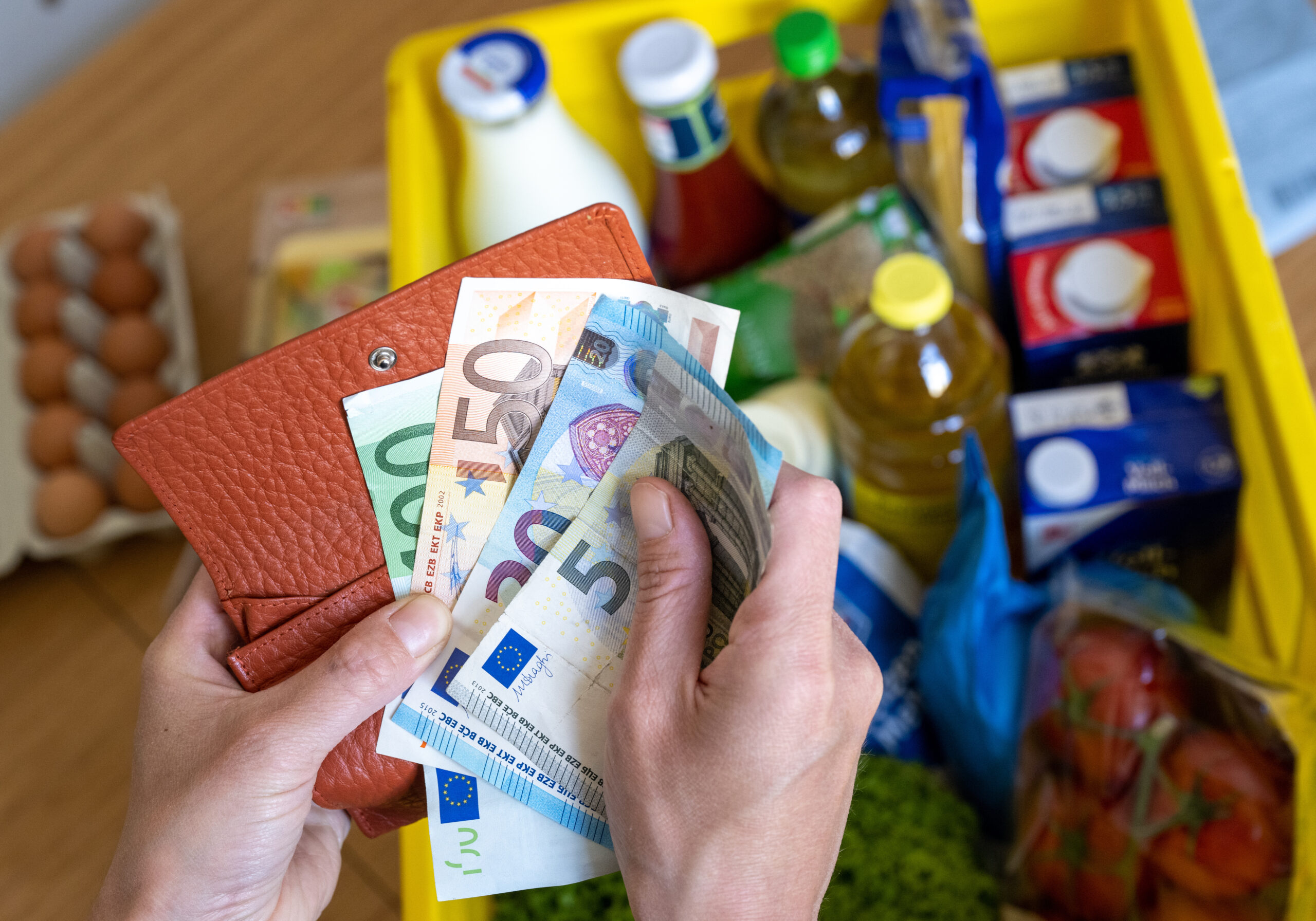Eine Einkaufskiste mit Lebensmitteln steht auf einem Küchentisch, während eine Frau Euro-Banknoten in den Händen hält.