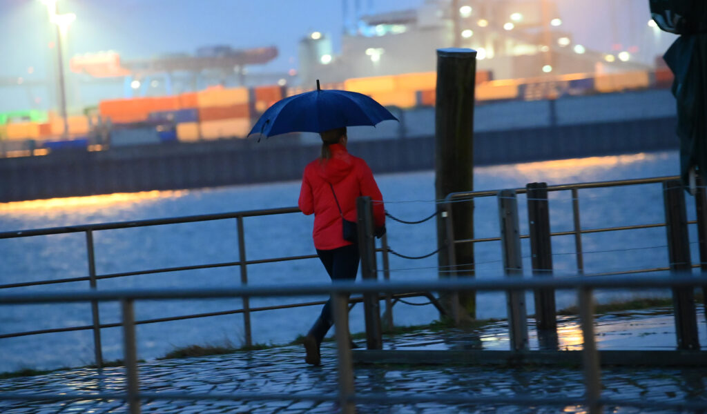 Eine Frau mit Schirm geht im Regen an der Elbe vor der Kulisse eines Containerterminals.