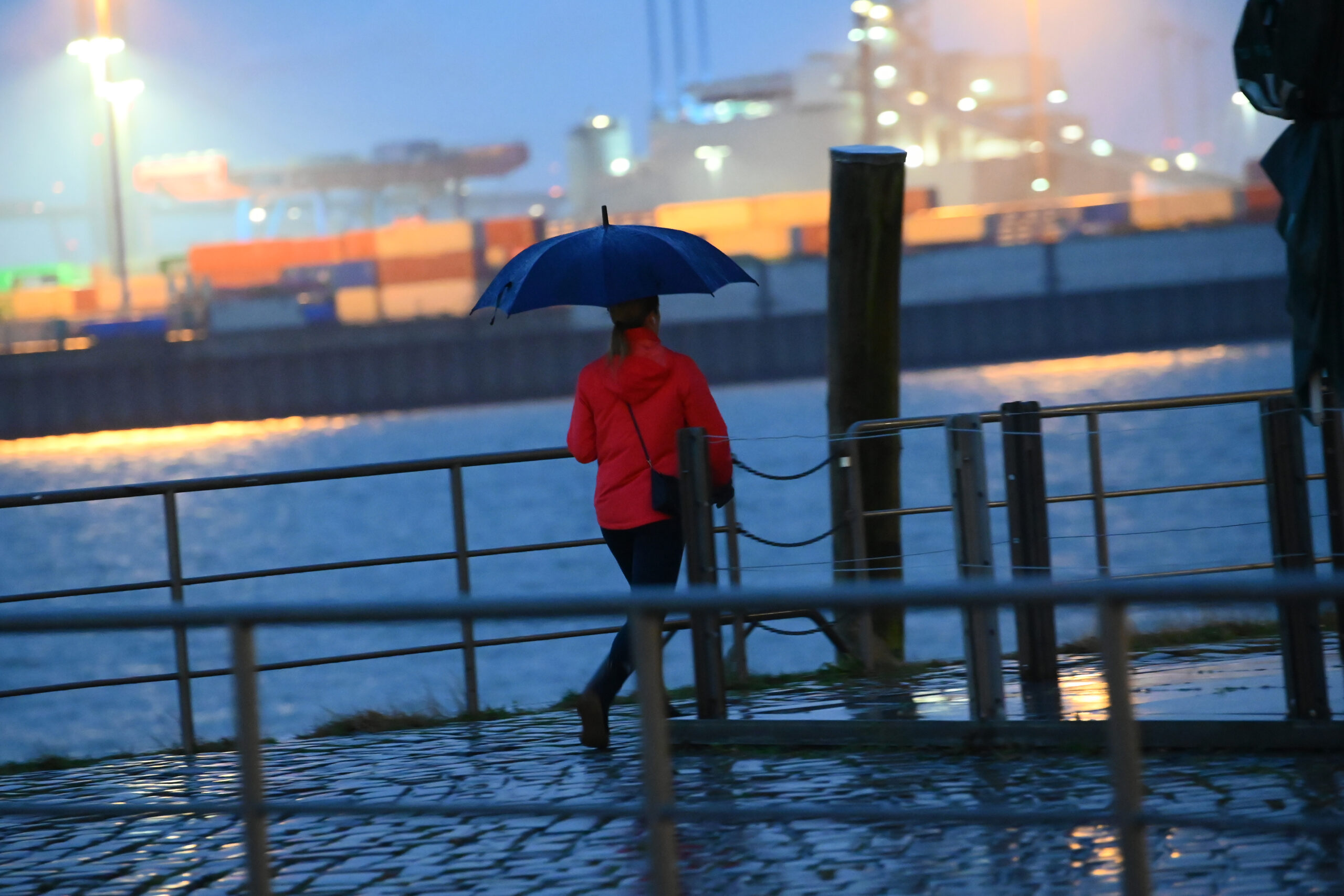 Eine Frau mit Schirm geht im Regen an der Elbe vor der Kulisse eines Containerterminals.