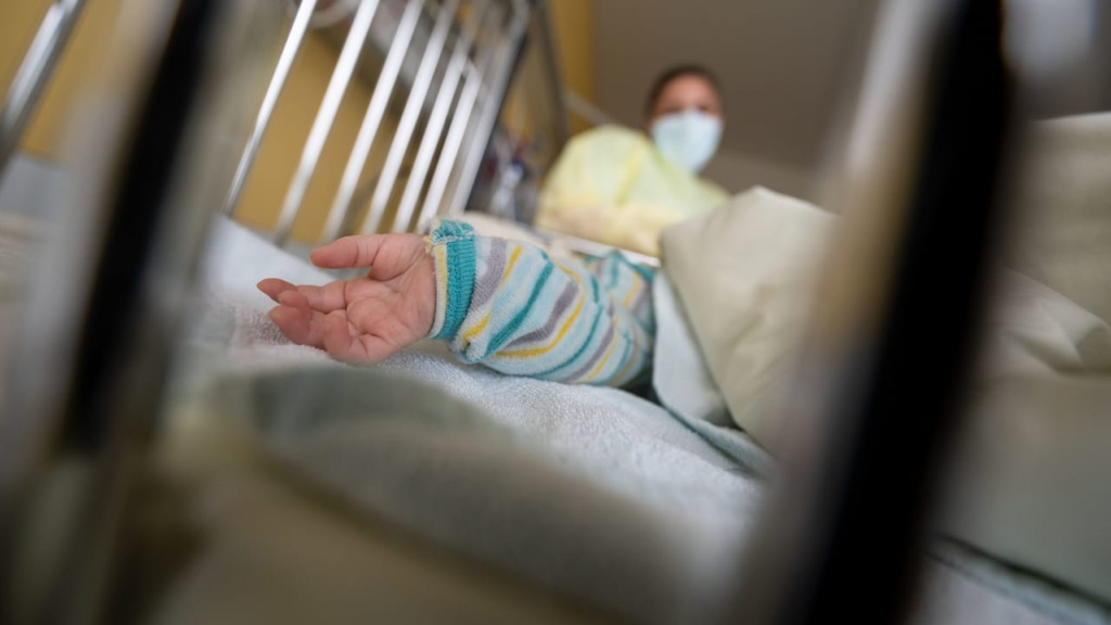 Eine Kinderhand guckt unter einer Decke hervor, dahinter eine Pflegerin im Schutzanzug