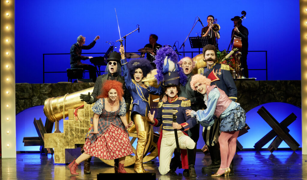 Das Ensemble in bunten Kostümen, im Hintergrund eine Band