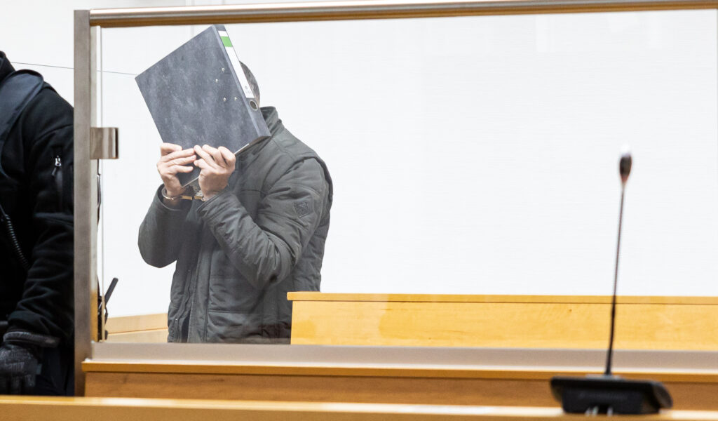 Ein Angeklagter mit Handschellen verdeckt sein Gesicht hinter einer Akte