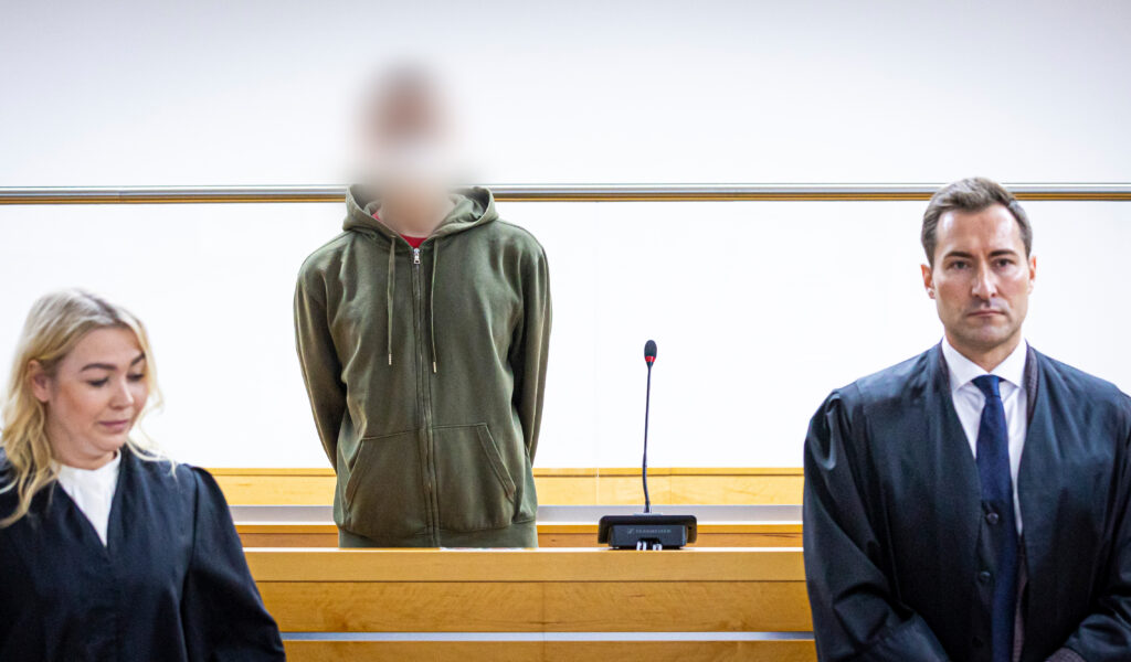 Ein Angeklagter in Olive-grüner Strickjacke steht zwischen seinen Anwälten