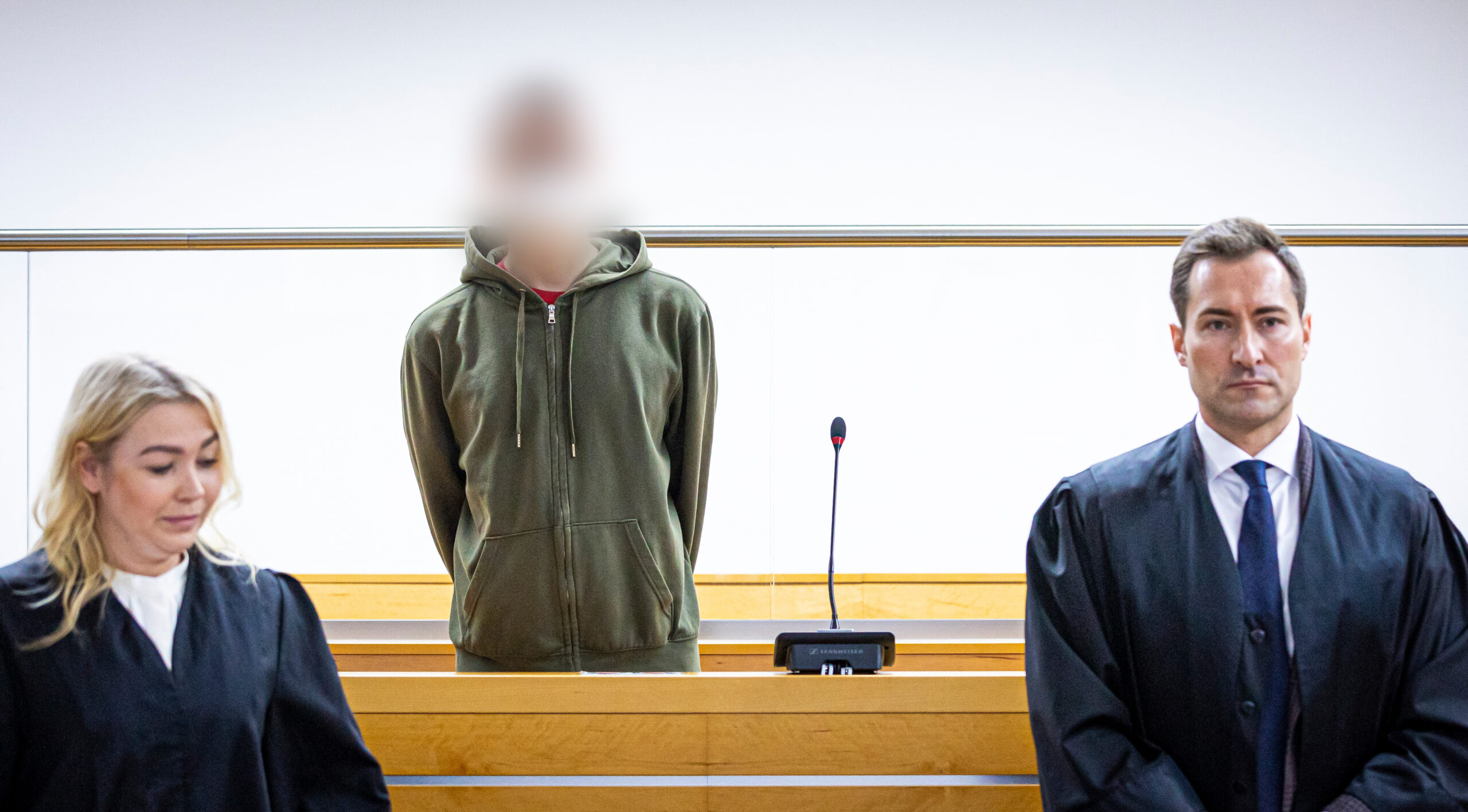 Ein Angeklagter in Olive-grüner Strickjacke steht zwischen seinen Anwälten