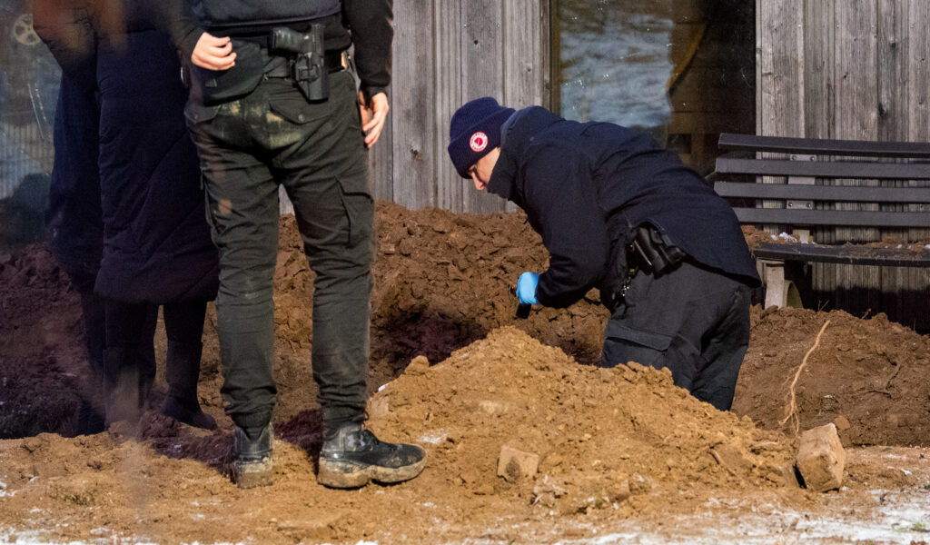 Ermittler der Polizei arbeiten hinter einem Mehrfamilienhaus in einem Garten an einem Erdloch. Dort wurde am Mittag eine vergrabene Leiche gefunden