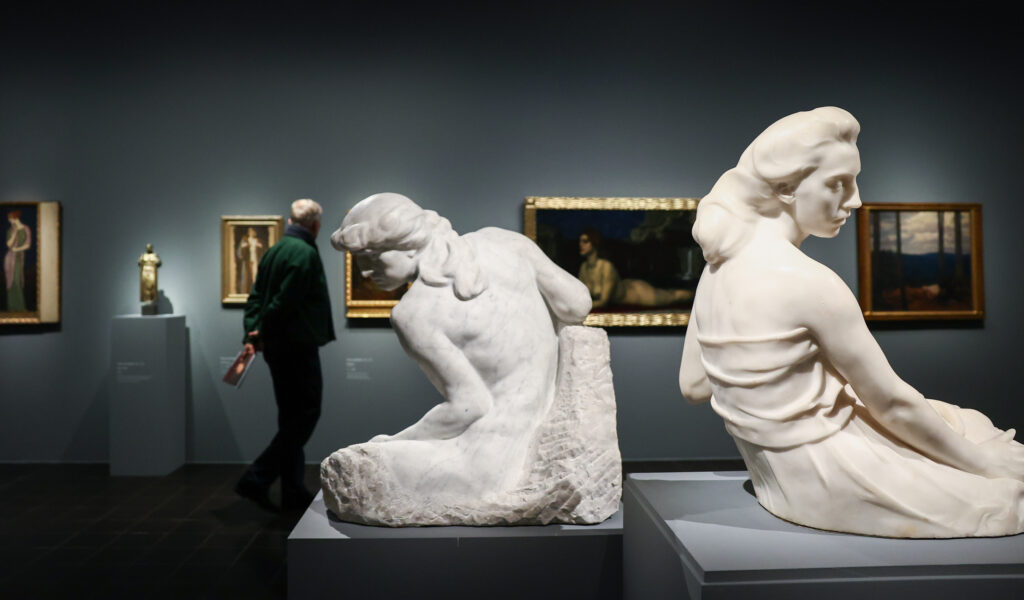 Im Vordergrund zwei Frauen-Skulpturen, im Hintergrund betrachtet ein Mann Gemälde an der Wand