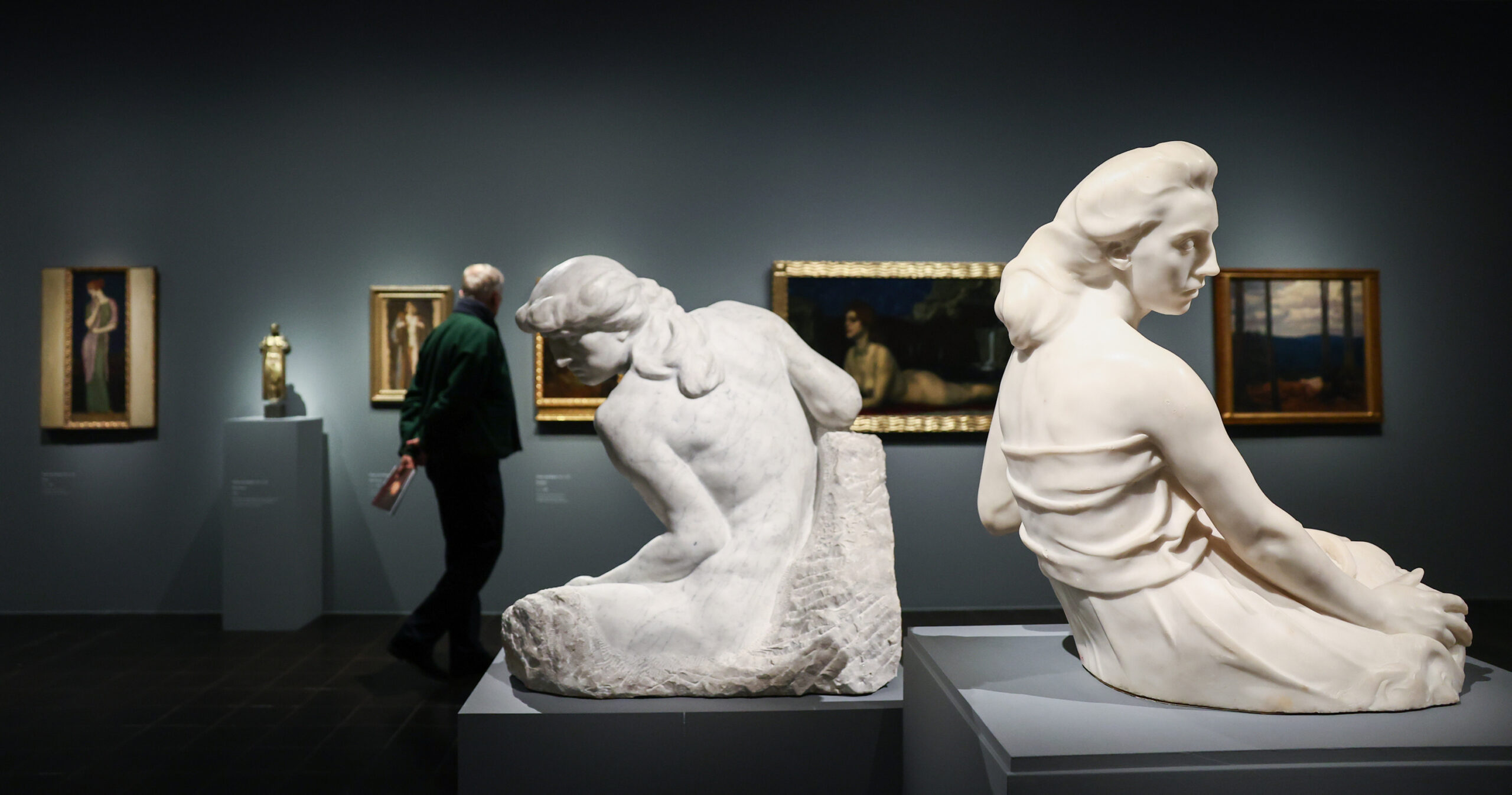 Im Vordergrund zwei Frauen-Skulpturen, im Hintergrund betrachtet ein Mann Gemälde an der Wand