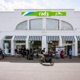 Mehrere Personen stehen vor einem Ladengeschäft mit dem Schriftzug 'rad3'. Ein Mann fährt mit einem Lastenrad los.