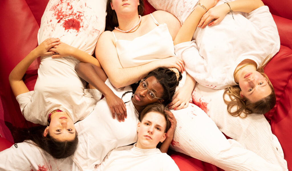 Fünf Schauspieler:innen liegen auf einer roten Matte, sie tragen weiße Kleidung mit blutigen Stellen