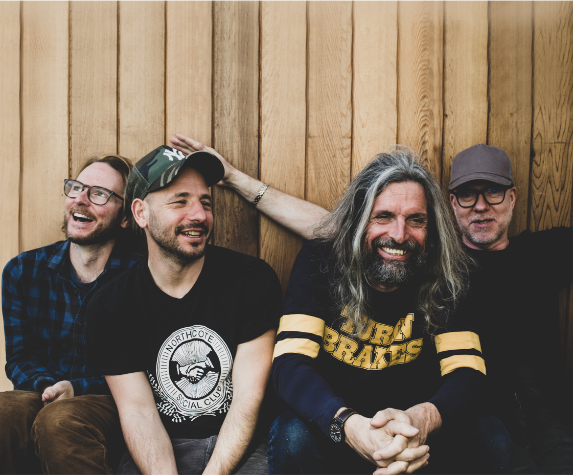 Die vier Musiker sitzen vor einer Holzwand und lächeln beziehungsweise lachen